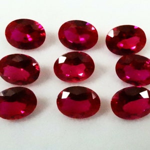 Puede incluir: Nueve piedras preciosas rojas oscuras de corte ovalado dispuestas sobre una superficie blanca. Las gemas tienen una superficie pulida y reflectante, mostrando su color y claridad vibrantes. Estas piedras son probablemente rubíes.