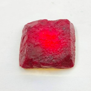 Può includere: Una gemma di rubino grezza e non tagliata, di colore rosso intenso. La superficie ha un aspetto ruvido e strutturato, e la pietra è illuminata dall'interno, creando un effetto luminoso. La gemma è di forma quadrata.