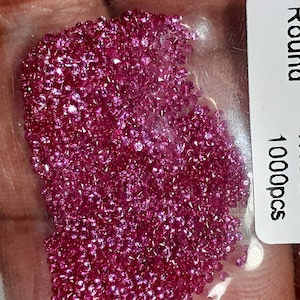 Op de afbeelding: Een doorzichtige plastic zak gevuld met talrijke kleine, ronde, helder roze edelstenen. De edelstenen zijn dicht opeengepakt, waardoor een sprankelend effect ontstaat. Een wit label met het woord "Round" en "1000pcs" is zichtbaar.