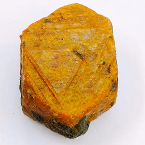 May include: A rough-cut, golden-yellow gemstone with a rectangular shape. The stone has a textured surface with visible lines and dark spots. The bottom edge is a darker, brownish-green color.