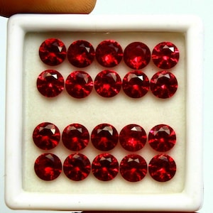 Könnte beinhalten: Sechzehn runde, facettierte, tiefrote Edelsteine ​​sind in einer weißen Schale angeordnet. Die Edelsteine ​​sind gleichmäßig in vier Reihen zu je vier Stück angeordnet, jede mit einer brillanten, reflektierenden Oberfläche. Die Schale steht vor einem hellen, neutralen Hintergrund.