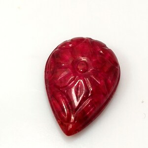 Puede incluir: Una piedra preciosa en forma de lágrima de un color rojo intenso y vibrante. La superficie de la piedra presenta un intrincado patrón floral tallado con un detalle circular central y pétalos radiantes, mostrando una textura pulida y translúcida.