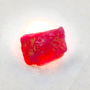 May include: A raw, unpolished gemstone with a vibrant, translucent neon pink and red hue. The stone features an irregular, angular shape with textured, pitted surfaces and natural inclusions visible throughout its crystalline structure.