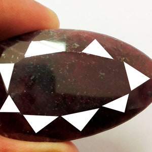 Può includere: Una gemma a forma di pera, di colore rosso scuro, con sfaccettature triangolari bianche. La gemma è tenuta tra le dita, mostrando la sua superficie lucida e il suo taglio unico. Il colore della gemma è un rosso intenso e ricco.