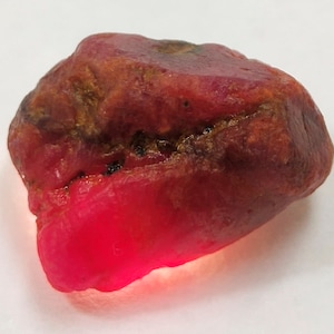 Op de afbeelding: Een ruwe robijn edelsteen met een dieprode kleur. De steen heeft een natuurlijke, onregelmatige vorm met een enigszins doorschijnend, gloeiend uiterlijk aan één kant. Het oppervlak vertoont kleur- en textuurvariaties.