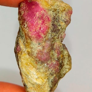 May include: A rough, unpolished pink ruby gemstone with a yellow-brown exterior. The ruby has a natural, raw appearance.