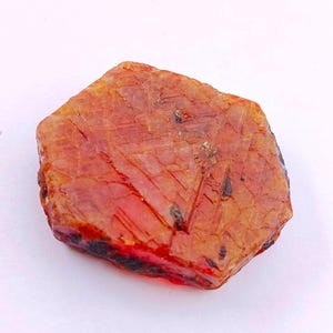 May include: A rough-cut, reddish-orange gemstone with a faceted surface. The stone has a hexagonal shape and displays a mix of light and dark areas, with some black inclusions. The overall appearance is natural and unpolished.