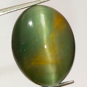 May include: An oval-shaped cabochon gemstone with a smooth, polished surface. The stone displays a translucent green colour with a distinct vertical band of light, known as chatoyancy, and a subtle golden-yellow internal inclusion pattern.