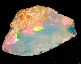 May include: A raw opal gemstone with a rough, textured surface. The opal displays a play of color, with flashes of pink, green, and blue within a translucent, milky white base. The top portion has a rough, tan-colored matrix.
