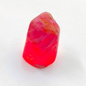 Peut inclure: Une pierre précieuse de rubis brut, d'un rouge profond. Le cristal naturel a une forme légèrement irrégulière et un aspect poli et translucide. La pierre est posée sur un fond blanc.