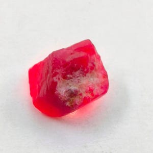 Può includere: Una gemma di rubino grezza e non tagliata. Il rubino è di un colore rosso vibrante con una superficie leggermente ruvida e strutturata. La gemma ha una forma naturale e irregolare ed è posta su uno sfondo bianco. Adatto per la creazione di gioielli.