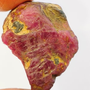 May include: A rough, red ruby gemstone with a dark red color and a rough, natural texture. The ruby is a deep red color with some brown and black inclusions.