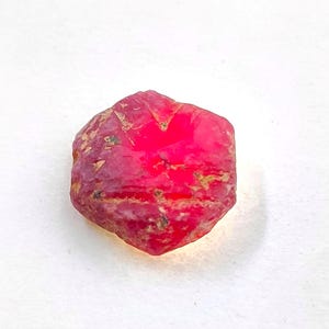 Pode incluir: Uma pedra preciosa de rubi bruta de forma hexagonal com uma cor vermelha vibrante e saturada. A superfície da pedra apresenta texturas naturais irregulares, pequenas inclusões e bordas facetadas que captam a luz de forma natural.