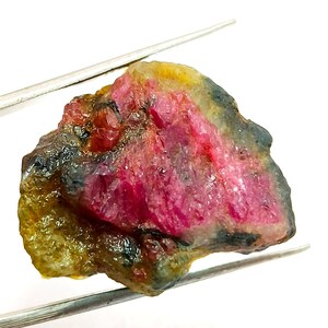 Puede incluir: Una gema en bruto con un vibrante centro rosa, que transiciona a tonos amarillos y verdes en los bordes. Sujeta por pinzas plateadas, la piedra muestra una textura natural y sin pulir. Una pieza única de mineral en bruto.