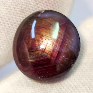 May include: A round, polished gemstone with a deep burgundy color and internal striations. The stone has a reflective surface with highlights of gold and purple. The gemstone is set against a white background.