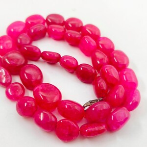 Könnte beinhalten: Eine Halskette aus polierten, oval geformten, pinkfarbenen Perlen. Die Perlen sind aufgereiht und bilden eine vollständige Schlaufe mit einem silberfarbenen Verschluss. Die Halskette wird vor einem weißen Hintergrund präsentiert.