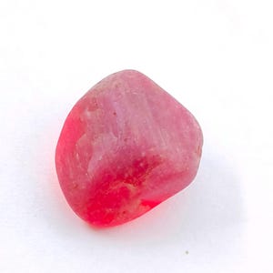 Könnte beinhalten: Ein glatter, abgerundeter, rosa Edelstein mit polierter Oberfläche. Der Stein hat eine lebendige, gesättigte Farbe, die von hellrosa bis tiefrosa reicht. Der Edelstein ist vor einem weißen Hintergrund platziert.
