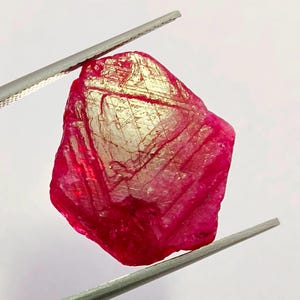 以下が含まれることがあります： 銀色のピンセットで保持された、未加工の鮮やかな赤色の宝石。 宝石は、反射性の金色の表面を持つ結晶構造をしています。 不規則な形状と深い色は、ジュエリーによく使用される貴重な石であるルビーを示唆しています。