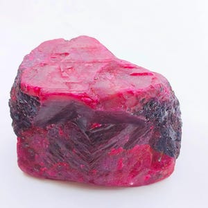 Puede incluir: Una gema de rubí en bruto, de color rojo intenso con bordes oscuros y texturizados. La piedra sin cortar muestra una estructura cristalina y una forma natural rugosa, mostrando su color vibrante y características geológicas únicas.