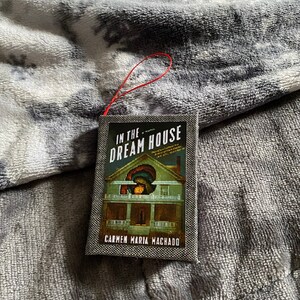 May include: A paperback book titled "In the Dream House" by Carmen Maria Machado. The cover features a dark, vintage-style illustration of a house with a woman looking out a window.