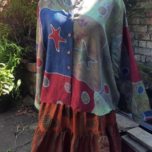 May include: A patterned shirt and tiered skirt outfit. The shirt has a colourful design with red stars and blue, green, and brown shapes. The skirt is a tiered, rust-coloured fabric. The outfit is displayed on a mannequin.