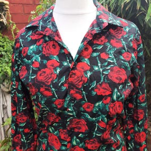 May include: A long-sleeved button-down shirt featuring a black background with a vibrant all-over print of red roses and green leaves. The shirt has a classic collar, a front button placket with black buttons, and is displayed on a white mannequin.