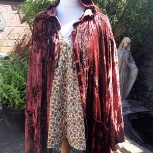 May include: A deep burgundy velvet hooded coat with pockets, worn over a floral print dress. The coat features a button closure and is displayed on a mannequin. The background includes greenery and a brick wall.