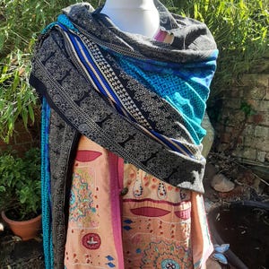 May include: A mannequin showcases a vibrant shawl and garment. The shawl has a black border with a white floral design, a blue stripe, and a turquoise centre. The garment beneath is peach with embroidered details and pink trim. Greenery forms the backdrop.