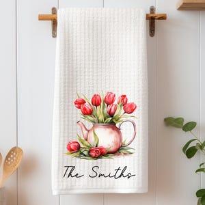 May include: White kitchen towel with a waffle weave texture, featuring a watercolor illustration of red tulips in a pink teapot. The text "The Smiths" is written in a script font below the floral design. The towel is hanging on a wooden rack.