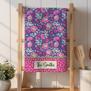 May include: A floral kitchen towel with a blue background and pink flowers, displayed on a wooden rack. The bottom border is pink with white polka dots, and a label reads "The Smiths".