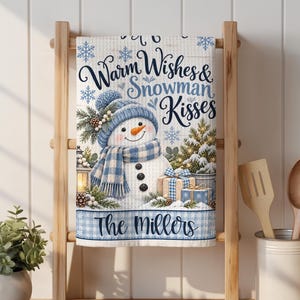 May include: A white kitchen towel featuring a snowman wearing a blue plaid hat and scarf, surrounded by snowflakes, a lantern, a pine tree, and gift boxes. The text reads Warm Wishes & Snowman Kisses and The Millers in blue script lettering.