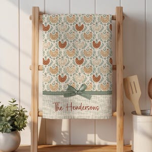 May include: A decorative kitchen towel with a repeating pattern of chickens and foliage in shades of brown, beige, and green. The towel is personalized with the text "The Hendersons" and features a green bow.