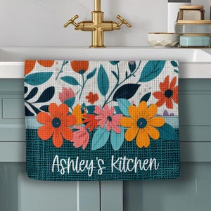 May include: A floral kitchen towel with a teal and white background. The towel features orange, pink, and yellow flowers with teal leaves. The words "Ashley's Kitchen" are printed in white script at the bottom.