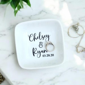 K&ouml;nnte beinhalten: Wei&szlig;es Keramik-Ringtablett mit den Namen "Chelsey & Ryan" und dem Datum "03.28.20" in schwarzer Schrift. Ein silberner Verlobungsring liegt auf dem Tablett. Das Tablett ist quadratisch mit abgerundeten Ecken.