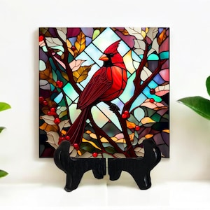 May include: A square decorative tile featuring a stained-glass style image of a red cardinal perched on a branch with red berries. The background is a colorful mosaic of blues, greens, and purples. The tile is displayed on a black wooden stand.