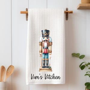 May include: White kitchen towel with a printed image of a nutcracker soldier. The nutcracker is in a military uniform with a black hat and gold accents. The text "Kim's Kitchen" is printed at the bottom of the towel.