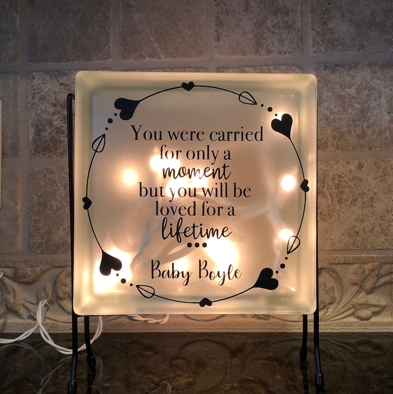 Puede incluir: Un bloque de vidrio cuadrado con el texto "You were carried for only a moment but you will be loved for a lifetime Baby Boyle" iluminado por peque&ntilde;as luces. El bloque est&aacute; decorado con un dise&ntilde;o de coraz&oacute;n y hojas negras y se encuentra sobre un soporte de metal negro.