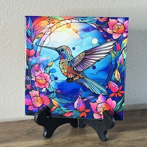 May include: A colorful stained glass style tile featuring a hummingbird with iridescent blue, green, and purple wings. The hummingbird is flying through a circular frame with flowers and leaves in shades of pink, red, and green.
