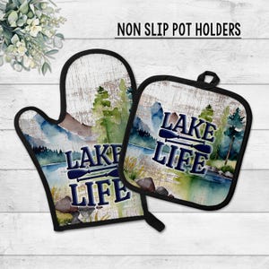 May include: Non-slip pot holders featuring a watercolor lake scene with the text "LAKE LIFE" and two crossed oars. The set includes an oven mitt and a square pot holder, both with black trim.