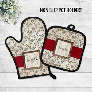 May include: A non-slip pot holder set featuring an oven mitt and a square pot holder. Both have a cream-colored background with a leaf pattern, a red stripe, and a framed initial with the family name. The set is trimmed in black.