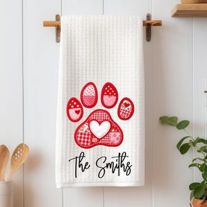 May include: White waffle-weave kitchen towel with a large, red paw print design. The paw print is made of various red and pink patterned fabrics, including hearts and polka dots. The text "The Smiths" is written in a script font below the paw print.