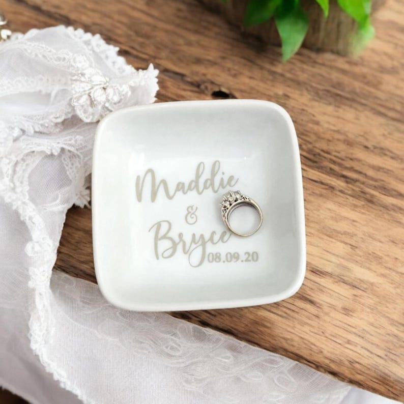 K&ouml;nnte beinhalten: Eine wei&szlig;e, quadratische Ringschale mit den Namen "Maddie & Bryce" und dem Datum "08.09.20" in Grau aufgedruckt. Ein silberner Ring mit dekorativer Fassung liegt in der Schale. Die Schale steht auf einer Holzoberfl&auml;che.