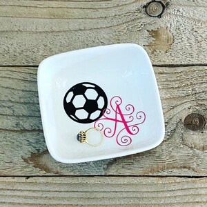 Puede incluir: Plato de cer&aacute;mica blanca para joyas con un dise&ntilde;o de bal&oacute;n de f&uacute;tbol blanco y negro y una letra A rosa con espirales. Un anillo de oro con una piedra azul est&aacute; sentado en el plato.
