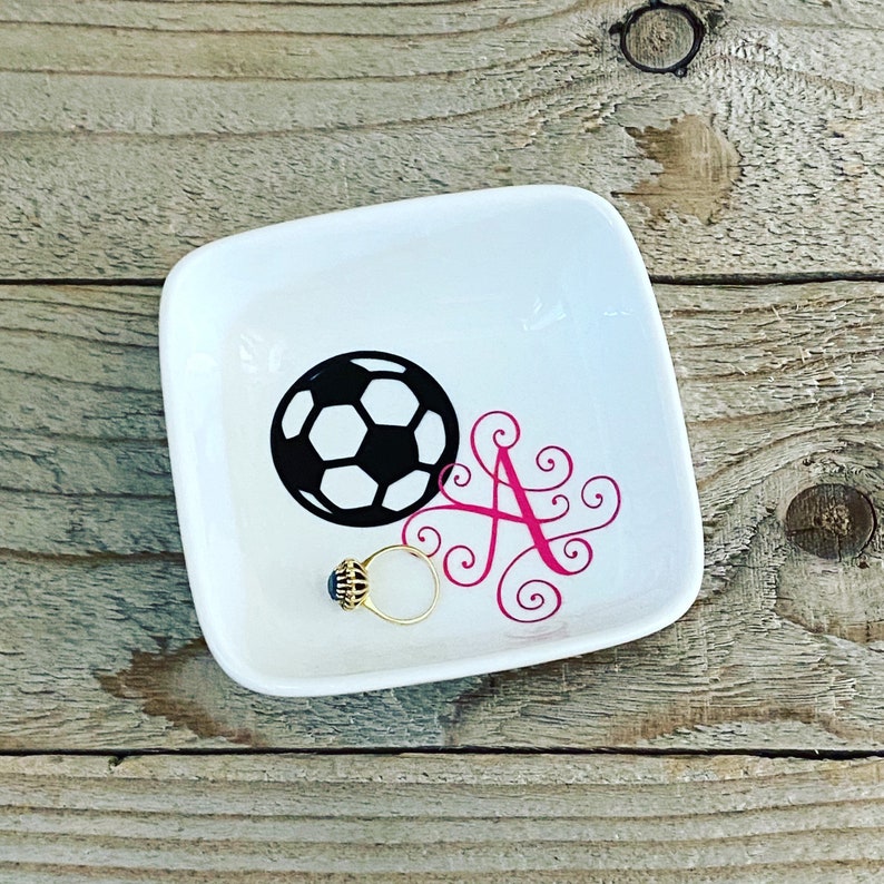 Puede incluir: Plato de cer&aacute;mica blanca para joyas con un dise&ntilde;o de bal&oacute;n de f&uacute;tbol blanco y negro y una letra A rosa con espirales. Un anillo de oro con una piedra azul est&aacute; sentado en el plato.