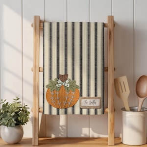 May include: A decorative kitchen towel with a black and cream striped pattern. The towel features an appliqued orange pumpkin with green leaves and a bow. A small rectangular tag below the pumpkin reads "The Smiths". The towel is displayed on a wooden ladder.