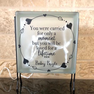 Puede incluir: Un bloque de vidrio cuadrado con el texto "You were carried for only a moment but you will be loved for a lifetime Baby Boyle" en escritura negra. El bloque est&aacute; decorado con un dise&ntilde;o de coraz&oacute;n y hoja y se apoya en un soporte de metal negro.