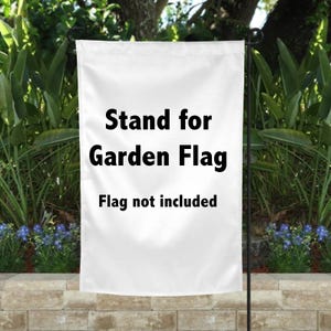 May include: A white garden flag stand with the text "Stand for Garden Flag. Flag not included" in black. The stand is displayed outdoors with greenery in the background.