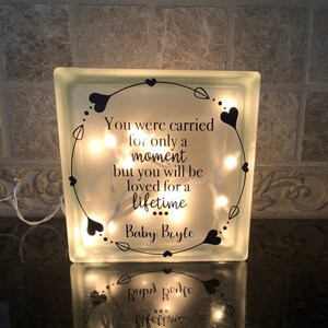 Puede incluir: Un bloque de vidrio transparente iluminado por luces de hadas. El bloque presenta un dise&ntilde;o de coraz&oacute;n y hojas negras y el texto: "You were carried for only a moment but you will be loved for a lifetime Baby Boyle."