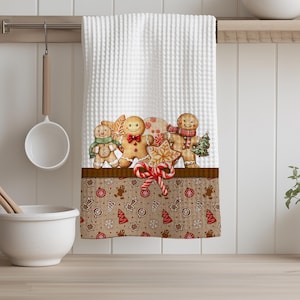 May include: White waffle-weave kitchen towel with a festive gingerbread design. The bottom border features a brown fabric with gingerbread men, stars, and Christmas trees. The towel is decorated with gingerbread men, candy canes, and a star-shaped cookie.