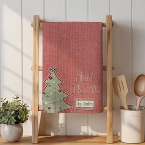 May include: A red kitchen towel with a stitched Christmas tree design and the words "MERRY CHRISTMAS" and "The Smiths." The towel is displayed on a wooden ladder rack, with kitchen utensils in a white container.